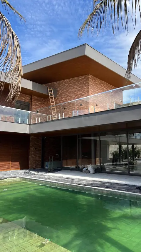 Casa moderna de dois pavimentos em fase de acabamento, com estrutura metálica aparente, revestimento em tijolinho e amplas esquadrias de vidro. A fachada conta com sacada com guarda-corpo de vidro e piscina de água esverdeada em primeiro plano.