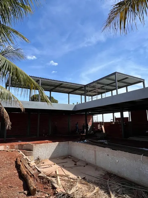 Construção de edifício de dois pavimentos em estrutura metálica, com pilares e vigas de aço aparentes e cobertura de telha metálica. O estágio atual mostra alvenaria de tijolos vermelhos no térreo e laje de concreto, em um canteiro de obras com materiais dispersos.