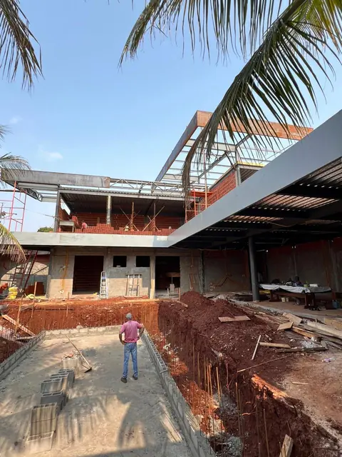 Construção de edifício de múltiplos pavimentos em estágio de estrutura mista, com alvenaria de tijolos vermelhos e vigas metálicas aparentes. Em primeiro plano, um operário observa o canteiro com solo escavado, blocos de concreto e ferragens expostas sob céu azul.