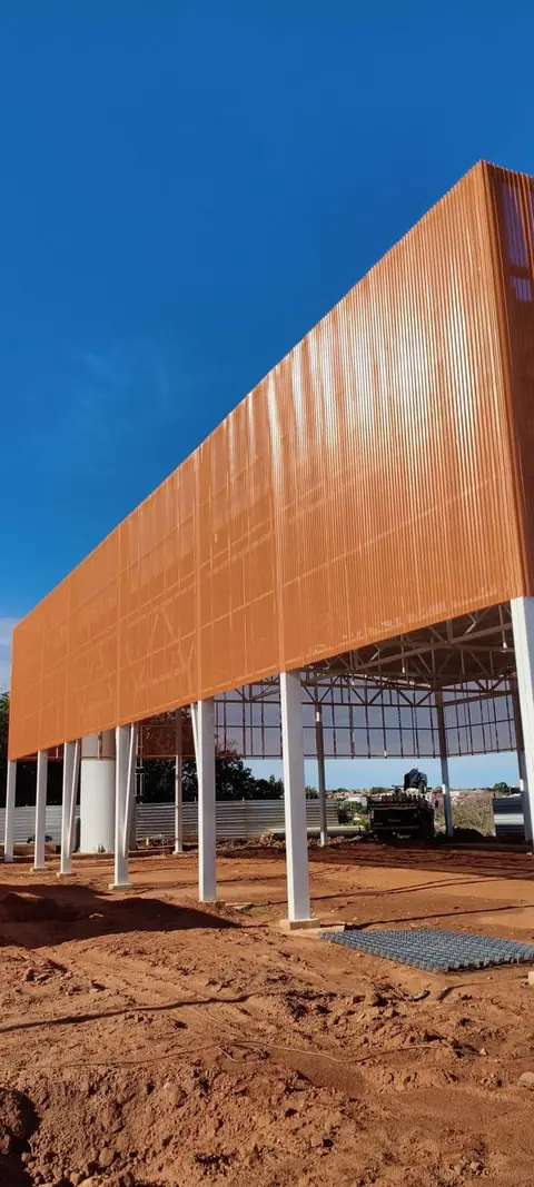 Estrutura metálica em estágio de montagem, com pilares brancos e um grande painel de fachada em chapa ondulada laranja, situada em um canteiro de obras de terra batida sob céu azul.