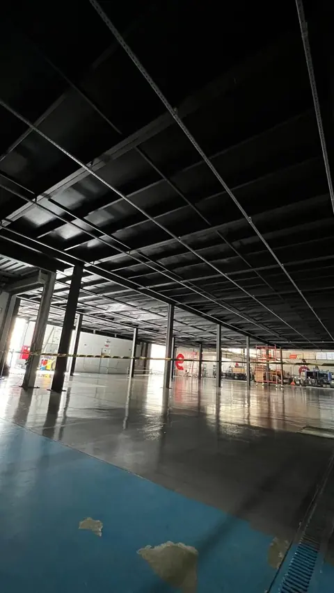 Interior de galpão industrial em fase de acabamento, com estrutura metálica aparente pintada de preto no teto e piso de resina azul e cinza polido. O ambiente amplo e vazio apresenta fitas de sinalização e equipamentos de construção ao fundo.