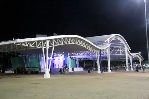 Estrutura metálica branca de grande porte com cobertura ondulada em estágio concluído, iluminada à noite. Abriga um palco com iluminação cênica azul e pessoas circulando em uma praça de terra batida.