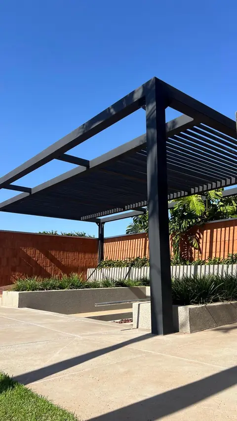 Estrutura moderna de pergolado de metal preto em fase de acabamento, com telhado ripado e sombras projetadas em piso de concreto sob céu azul.